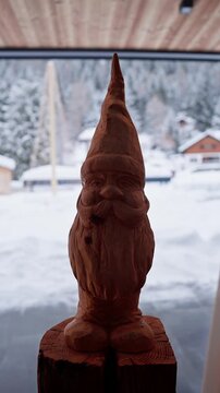 Hand carved wooden gnome stands on a split log in an alpine chalet, snowy fir trees and chalet rooftops beyond, winter daytime, subtle camera shifts, soft backlight.
