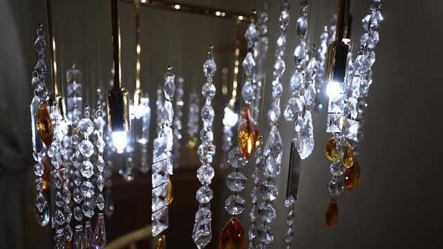 Interior chandelier shows gold rods, clear prisms, amber teardrops, and small LED bulbs in an Alpine chalet. Strands sway as refracted light sparkles in a dim evening.