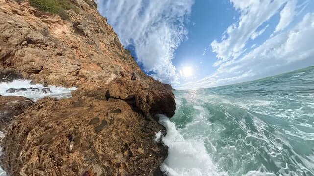 Man on a rock dodging the waves. A dramatic side-angle view of waves splashing high against coastal rocks, with sunlight streaming through scattered clouds over the shimmering sea