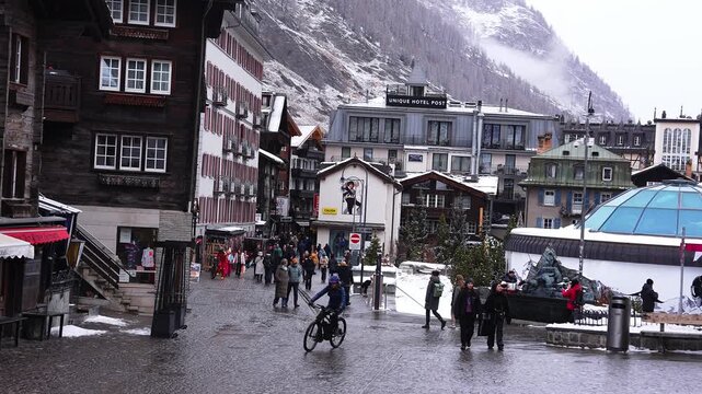 Wide street view in Zermatt shows cobbled plaza, UNIQUE HOTEL POST, blue glass dome, fountain, chalets, and hotels as shoppers, tourists, and a cyclist move in cool winter light.