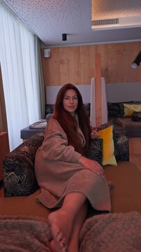 Red haired woman reclines on a sofa in a Zermatt spa lounge as she subtly shifts gaze and hand. Wood panels, linear fireplace, sheer curtains, and yellow pillows.