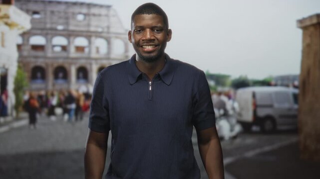 Man clapping hands and smiling before the colosseum building with tourists, scooters and a van on the cobbled street; joy travel.