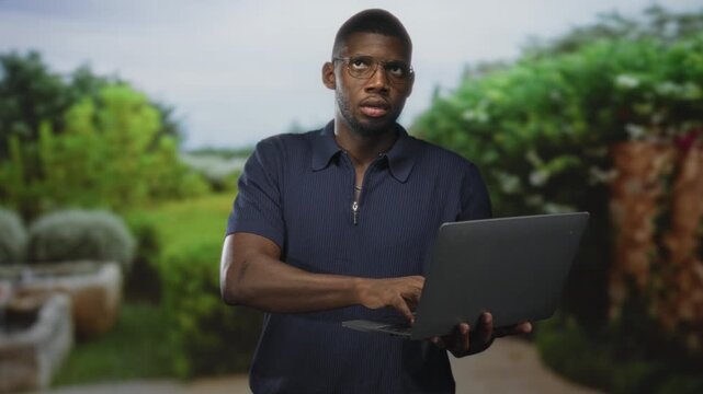 Man holding laptop and typing with right hand while looking at screen in studio setting with blurred plants; focused productivity.