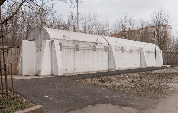 White Quonset Hut Shelter With Reinforced Concrete Walls And Metal Door In Rural Setting Amid Bare Trees And Overcast Sky