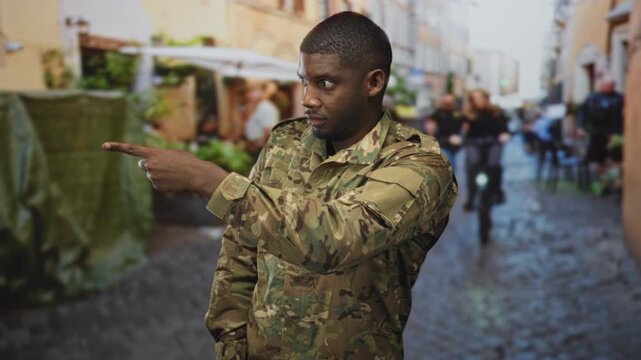 Soldier man pointing finger in camouflage uniform on a crowded cobblestone street while directing traffic and people; duty focus.