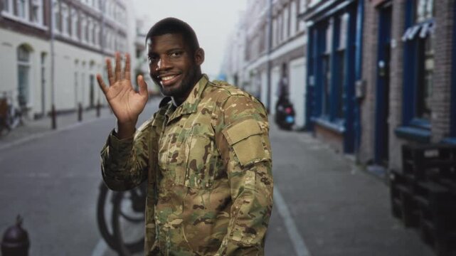Soldier man in camouflage uniform waving right hand on a narrow urban street with bicycle and shopfronts; pride duty.