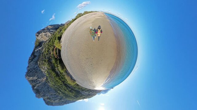 A tiny planet aerial view of a couple walking along a curved sandy shore, surrounded by turquoise water, lush green cliffs, and a vibrant blue sky