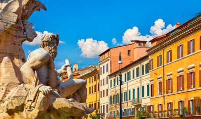 Marble sculpture of the Ganges River God at the Fountain of the Four Rivers in Rome, Italy. Historic Italian architecture and colorful buildings under a bright blue sky.