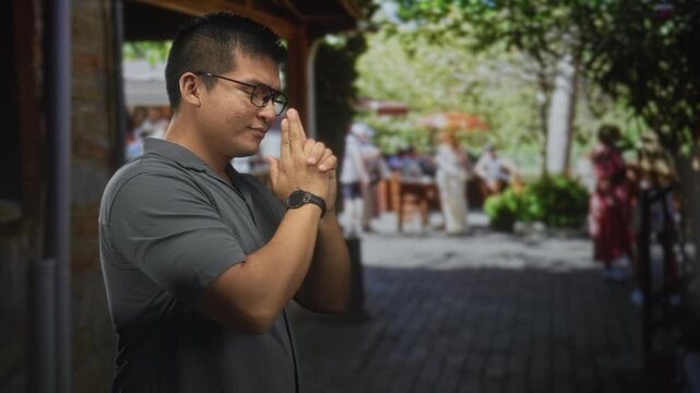 Man making finger gun pointing gesture with bare hands, wearing glasses and a wristwatch in a busy street cafe with blurred diners and trees; playful.