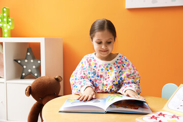 Smiling little girl reading book at table near orange wall indoors