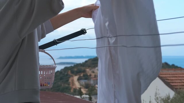 Housewife hanging fresh laundry on clothesline. Woman hanging clean white bed sheets on a clothesline with a basket full of clothespins, performing household chores with a beautiful sea view