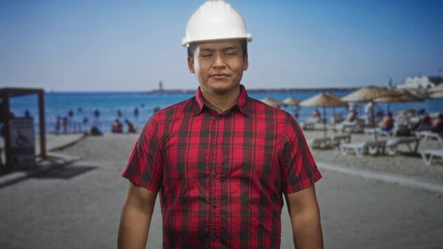 Man in white hardhat and red plaid shirt raises fist at a sunny beach with umbrellas and sunbeds, showing casual forearm; professional confidence.