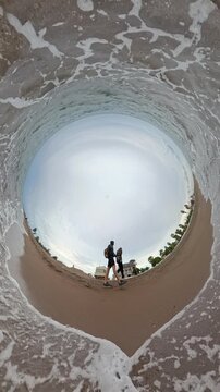Vertical video. Two people stroll by the edge of the water, captured with a Tiny Planet effect that distorts the beach and foam into a circular and dynamic spherical view. Overcast skies and cool weat