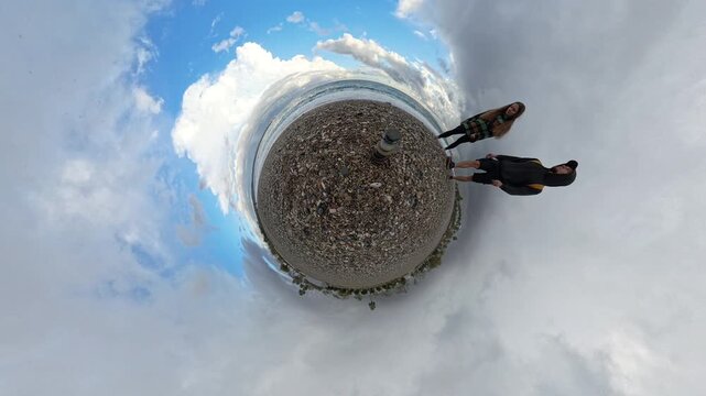 Tiny Planet view of a pebble beach featuring a balanced stone stack at the center, with a couple walking nearby under a cloudy, circular sky