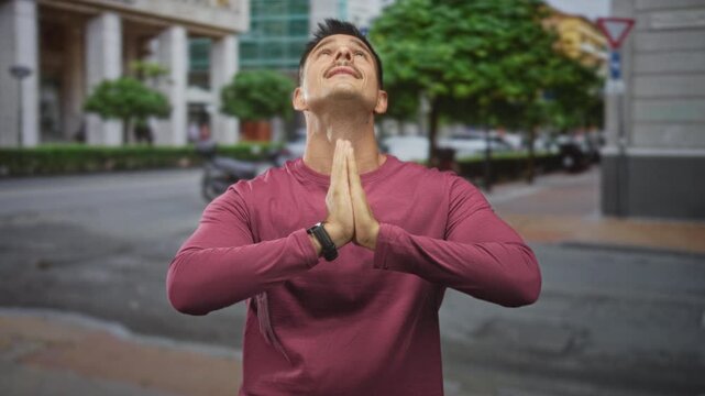 Man pressing hands in prayer gesture on a city street in front of a building, head tilted up showing moustache; gratitude reflection contemplation.