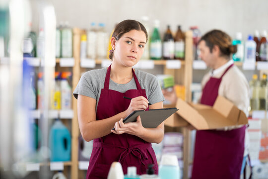 Girl seller, with tablet in hands, make inventory in store, man employee in background. Seller counts number of liquid powder bottles, records data in report, write down availability of goods in stock
