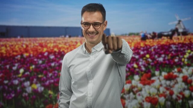 Man with moustache in striped shirt points finger toward camera in studio backdrop of tulip field and windmill; confidence invitation.