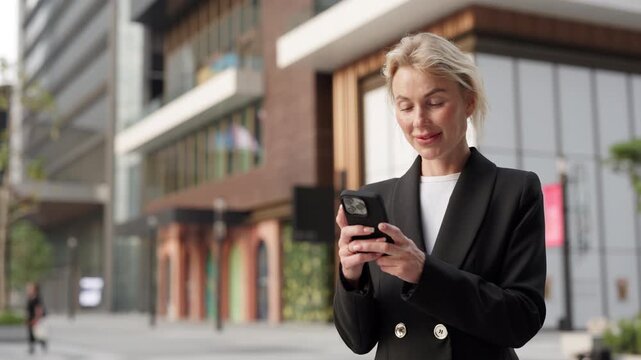 Portrait of happy businesswoman texts on smartphone device in downtown block. Female company CEO answers important message via cellphone standing near office building in city