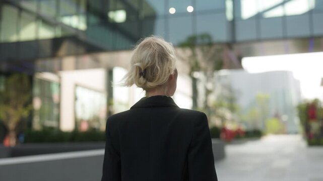Confident businesswoman in black jacket walks along downtown street backside view. Lady leader with blonde hair strolls alone in shady urban park horizontal shot. Corporate career