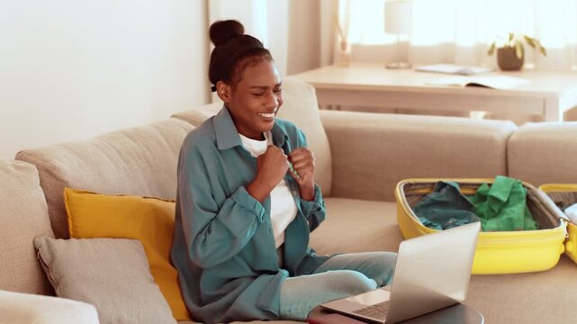 A woman with dark skin is sitting on a light-colored couch, smiling and engaging with a laptop. A suitcase is open nearby, showing colorful clothing, and sunlight fills the room.