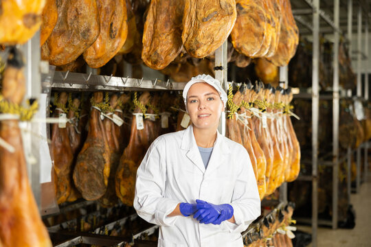 Girl employee in white uniform, bathrobe and hat, stands in workshop for production of ham. Working moments, close-knit team, private enterprise