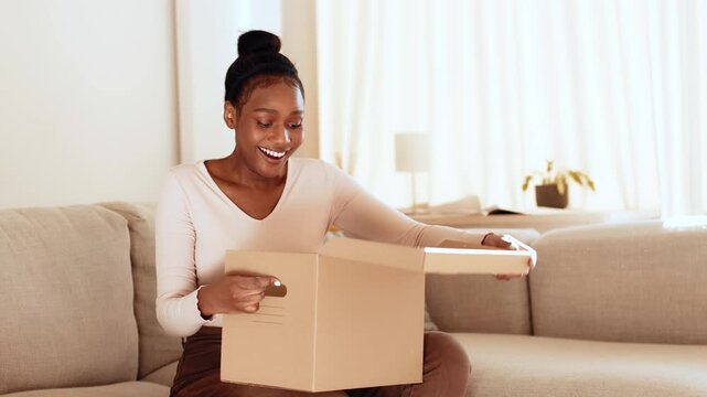 A woman with dark skin sits on a sofa in a well-lit living room. She is focused on opening a cardboard box in front of her. The environment is warm and inviting, filled with natural light.