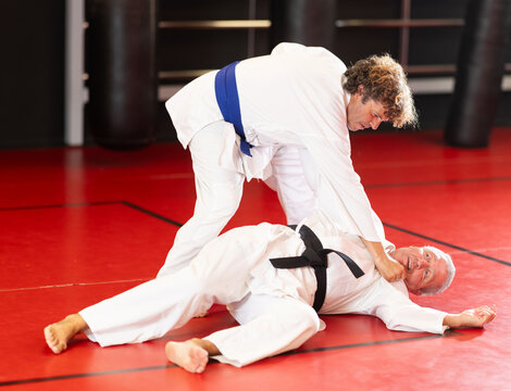 Concentrated blue-belt middle-aged aikido fighter in white gi honing fighting techniques during training kumite with older male partner in dojo. High quality photo