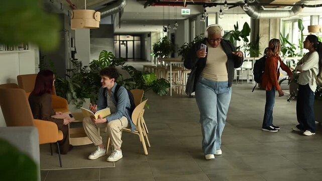 Joyful plus size student dancing with headphones while walking through a busy university campus common area