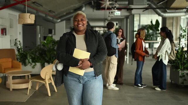 Confident african american female student standing and smiling in a busy university hallway with classmates in the background