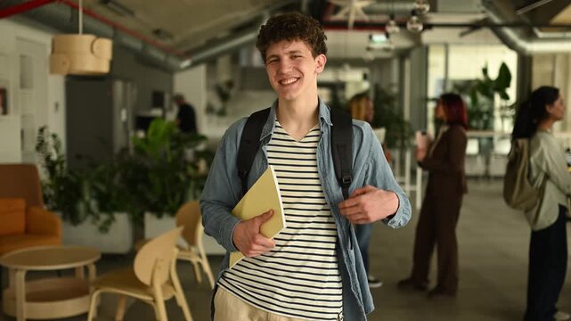 Portrait of a happy male college student standing in a modern university hallway and smiling at the camera