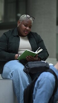 Young black woman sitting on a university campus bench reading a book aloud and memorizing notes for her final exams