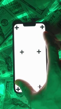 Vertical video. Top-View Shot of a Hand Performing a Zoom Gesture on a Smartphone with a Blank White Screen Mockup and Tracking Markers, Set Against a Green-Glowing Background of US Hundred-Dollar Bil