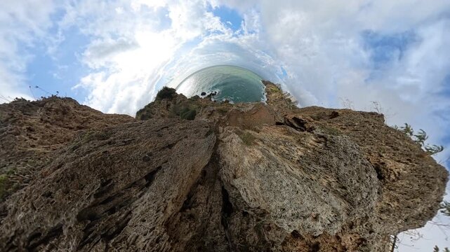 "Tiny planet" effect showcasing dramatic cliffside rocks curving toward the ocean, with a bright sun and scattered clouds creating a surreal panoramic view
