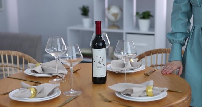 Beautiful place setting. Smiling woman in dress putting bottle of wine onto table at home, camera moving down and forward