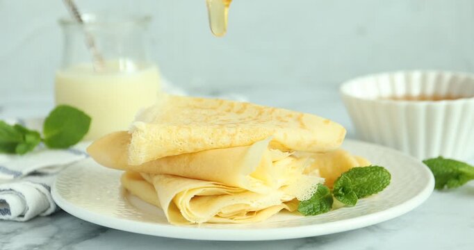 Woman dripping honey onto tasty crepes at white marble table, closeup