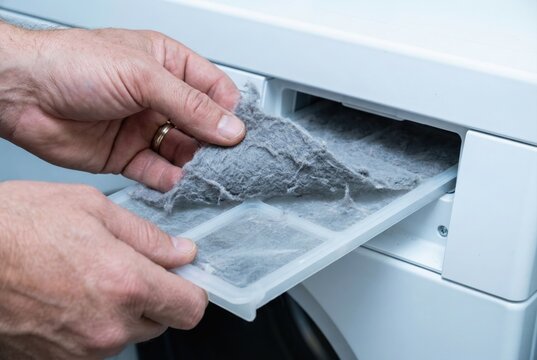 Hands peeling a thick layer of grey lint dust off a white tumble dryer filter screen