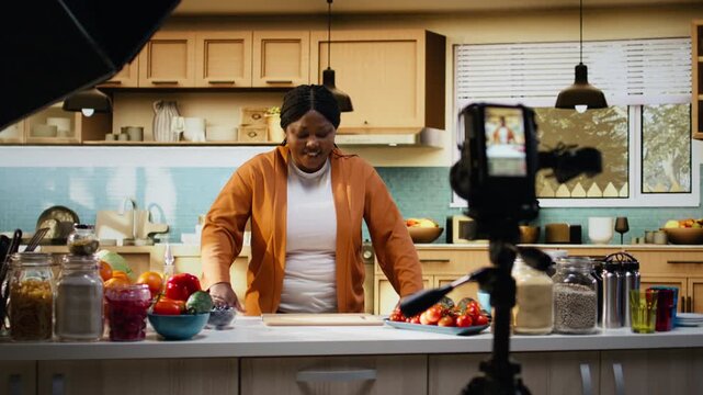 Behind the scenes of black woman recording meal prep vlog with tripod in the kitchen, presenting cooking process for online social media audience. Female influencer and content creator.