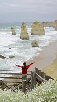 Traveler girl stands at the viewpoint spreading her hands and admiring powerful waves crashing against the limestone stacks of the Twelve Apostles on the Great Ocean Road, Victoria, Australia