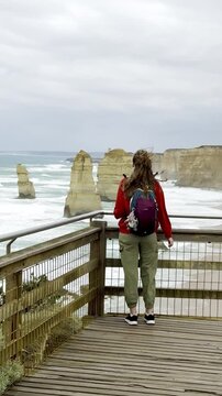 Traveler girl stands at the viewpoint and admires powerful waves crashing against the limestone stacks of the Twelve Apostles on the Great Ocean Road, Victoria, Australia