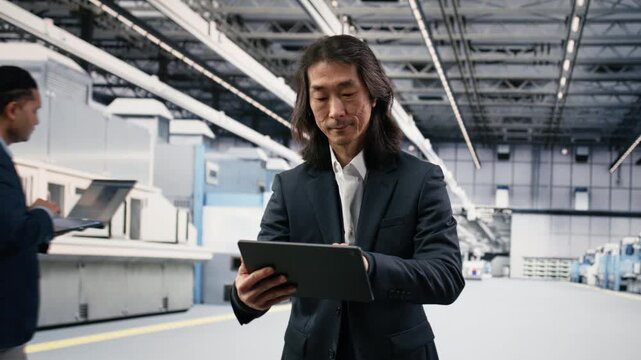 Microchip factory senior manager testing silicon wafer batches with tablet. Asian man in manufacturing plant monitors semiconductor machinery readouts with device, camera B