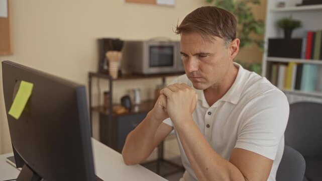 Man clasping hands at a computer monitor in a building office, seated at a desk wearing a white polo shirt and looking down; quiet concentration.
