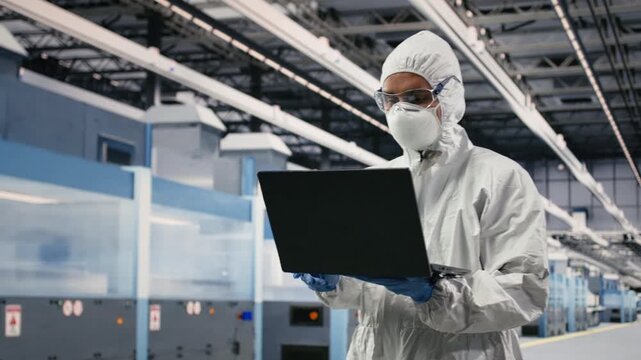 Scientist in microchip factory cleanroom uses laptop to improve integrated circuits design. Employee in bunny suit equipment uses notebook to supervise semiconductors manufacturing machinery, camera B