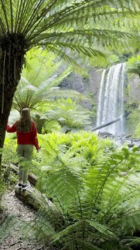 Girl adventurer stands next to tree fern and admires the powerful Hopetoun Falls, located in Great Otway National Park, Victoria, Australia. Stunning waterfalls in the Otways region