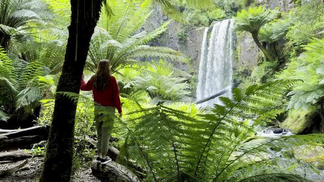 Girl adventurer stands next to tree fern and admires the powerful Hopetoun Falls, located in Great Otway National Park, Victoria, Australia. Stunning waterfalls in the Otways region