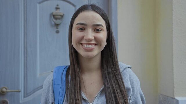 Young hispanic woman student wearing gray hoodie with blue backpack smiles at camera showing teeth and dimples at a building entrance doorway; optimism new start.