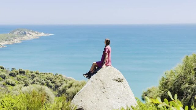 Girl adventurer sits on a large rock with her arms spread, admiring the stunning view over Fairy Cove and the turquoise ocean in Wilsons Promontory National Park, Victoria, Australia
