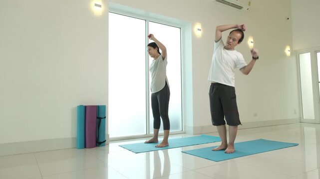 Man and woman practicing standing side stretch yoga pose at home, improving flexibility, spinal mobility and posture, reducing tension and enhancing balance in bright minimalist indoor fitness space