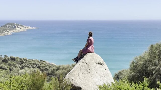Girl adventurer sits on a large rock with her arms spread, admiring the stunning view over Fairy Cove and the turquoise ocean in Wilsons Promontory National Park, Victoria, Australia