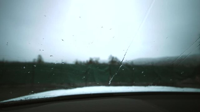Windshield wipers sweeping rainy countryside horizon, dashboard foreground, droplets streaking across glass, distant oil field silhouettes with pumpjacks and pipelines, overcast sky, muted