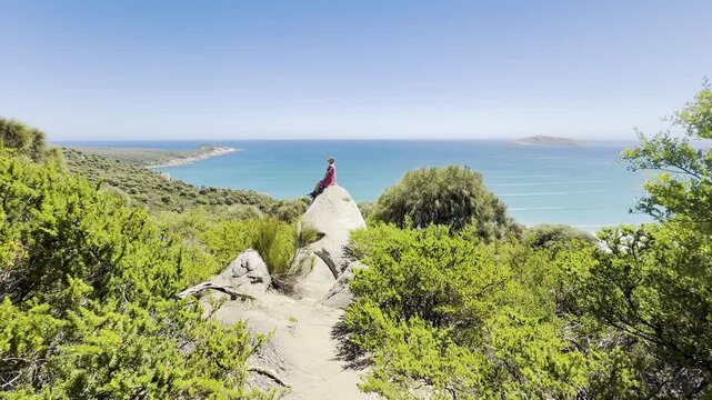 Girl adventurer sits on a large rock with her arms spread, admiring the stunning view over Fairy Cove and the turquoise ocean in Wilsons Promontory National Park, Victoria, Australia
